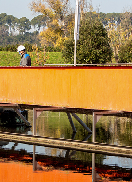 una vasca della sorgente del depuratore di roma nord con un ponte arancione in ferro su cui lavorano degli operai  acea infrastructure e gli alberi sullo sfondo